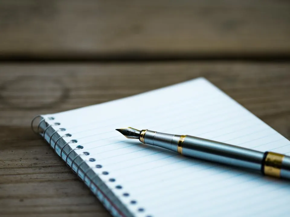 A silver fountain pen resting on a blank lined spiral notebook on a wooden table