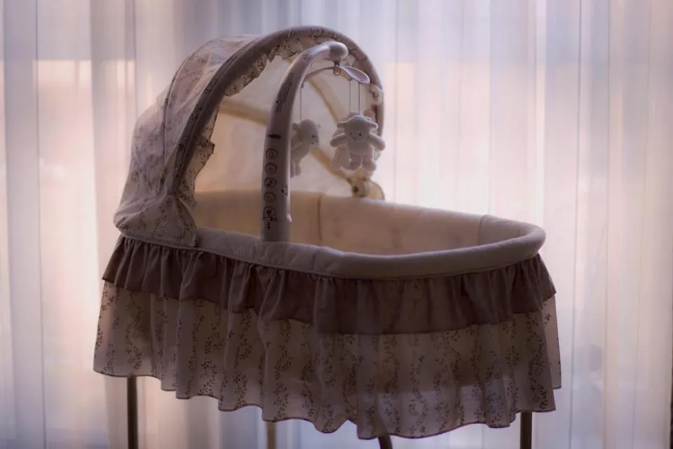 A ruffled baby bassinet with a canopy and hanging toys in front of sheer curtains