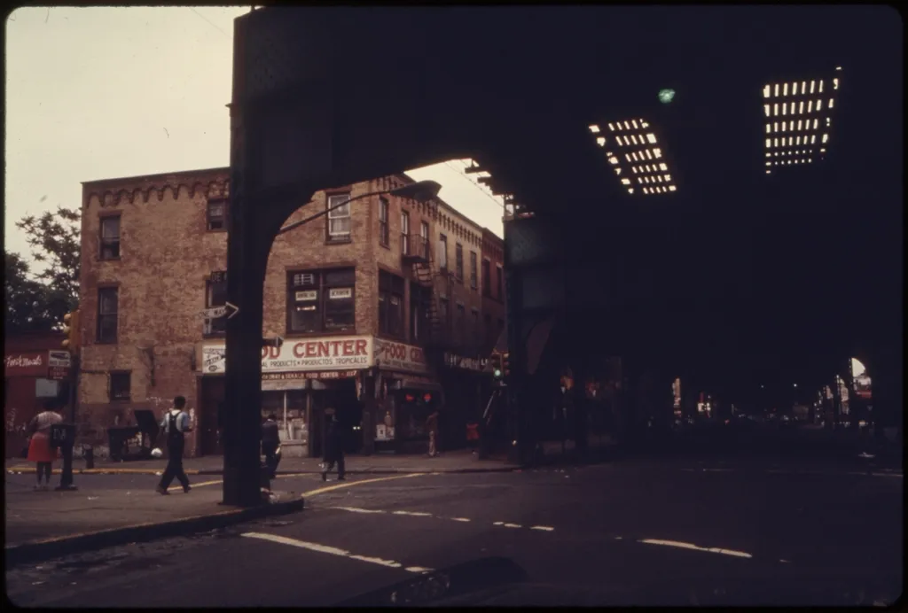 A city street with storefronts in the shadow of elevated train tracks in New York