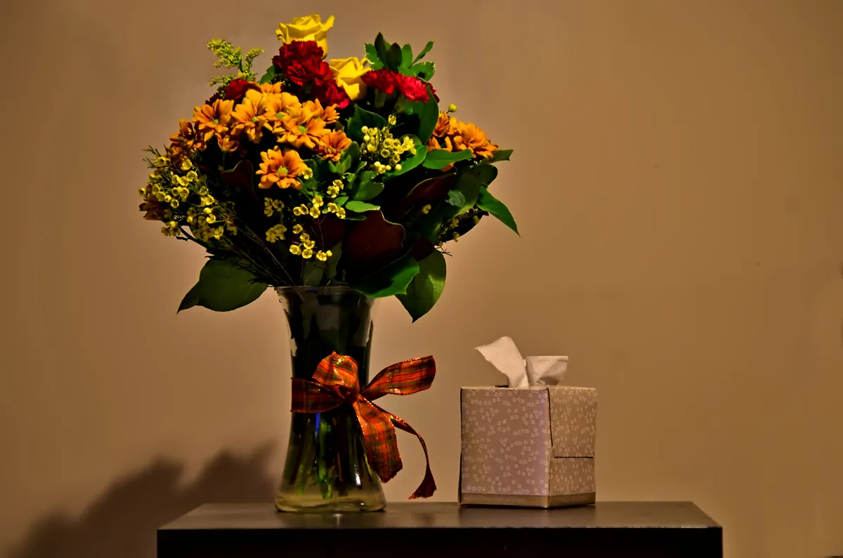 A vase of orange and red flowers beside a box of tissues on a dark shelf
