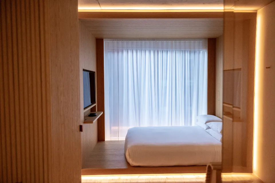 A minimalist hotel room with a low white bed, wood paneling and a curtained window