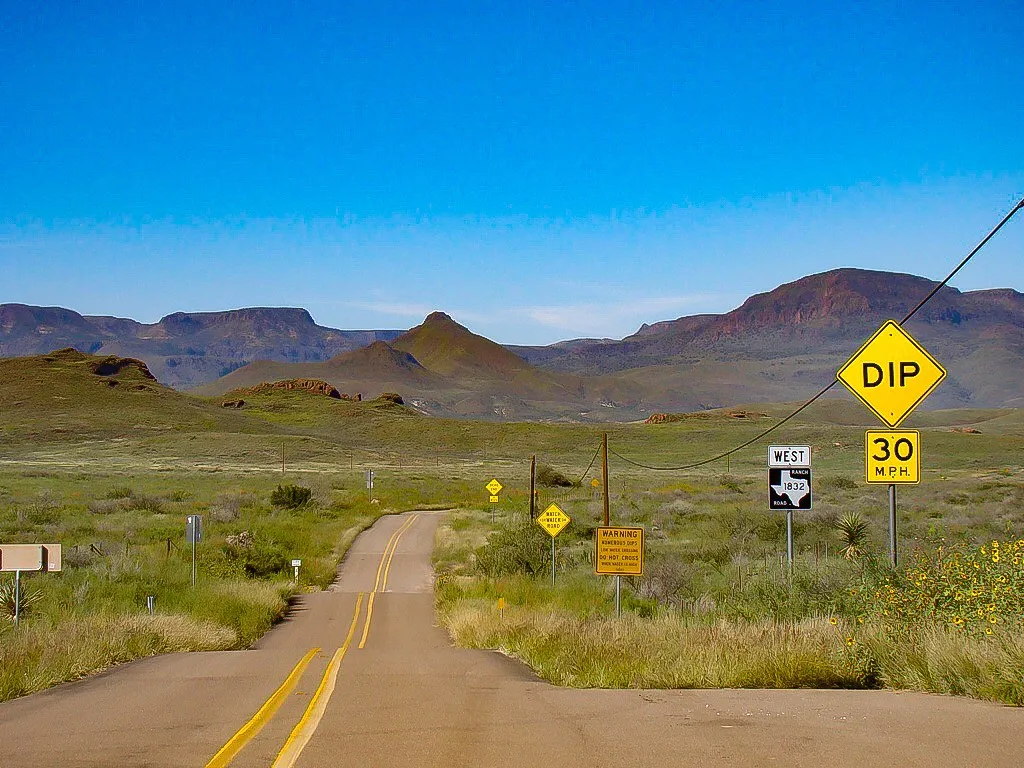 A two-lane Texas highway with a yellow dip warning sign and mesas in the distance