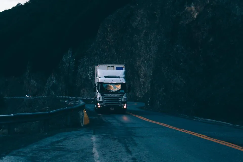 A white box truck with headlights on rounding a curve on a dark mountain road at dusk