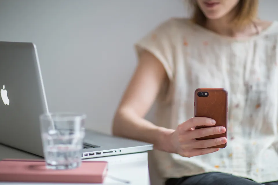 A woman's hand holding a smartphone beside a laptop and a glass of water on a desk