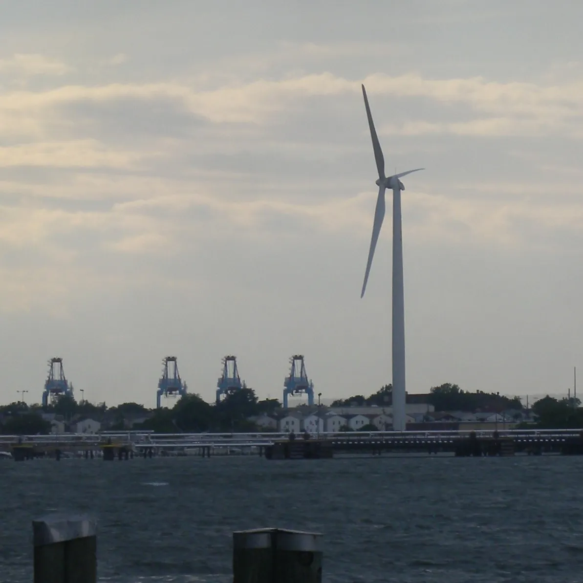 Container cranes on the New Jersey waterfront in Bayonne beside a tall wind turbine under gray sky