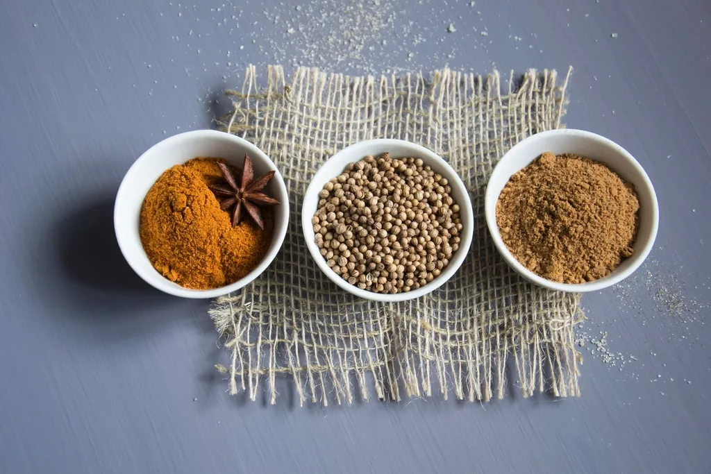 Three white bowls of ground spices and whole seeds on a square of burlap