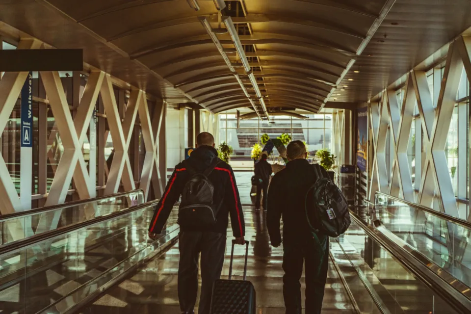 Two travelers seen from behind on an airport moving walkway pulling a rolling bag
