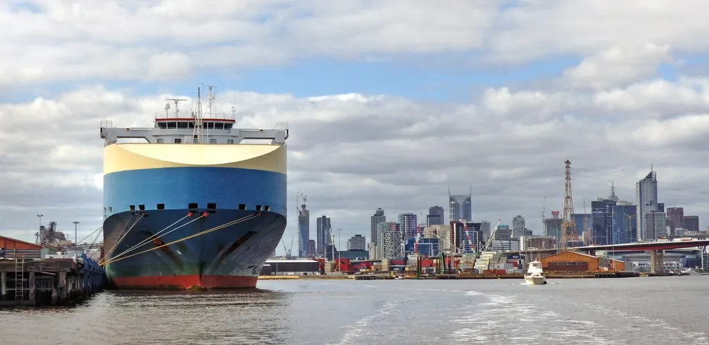 A car carrier ship docked at a port with a city skyline in the distance