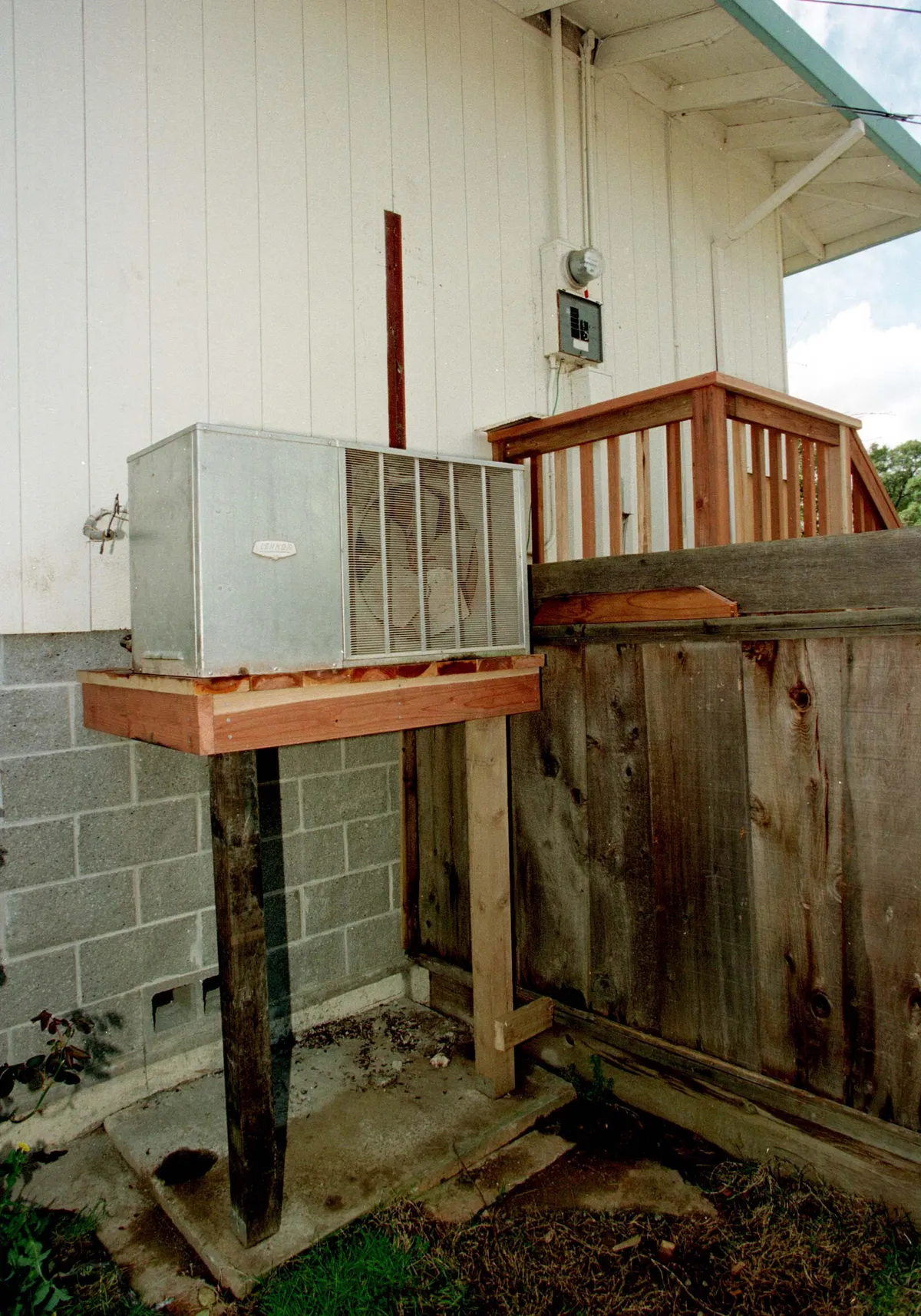 An outdoor air conditioning condenser unit mounted on a wooden platform beside a house