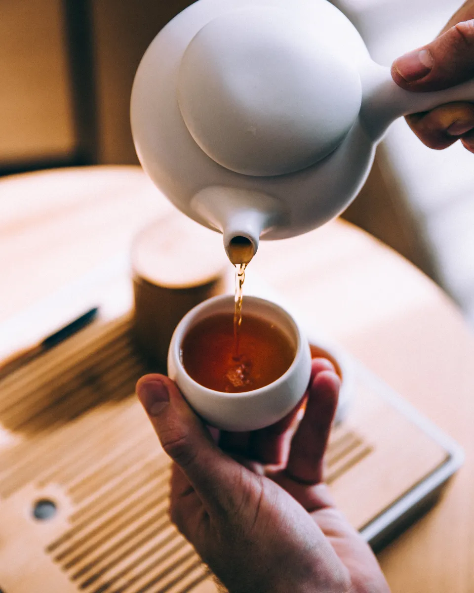 Hands pouring tea from a white teapot into a small cup