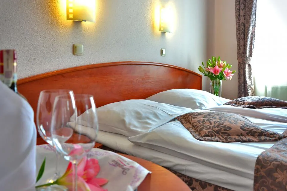 A made hotel bed with white linens, pink flowers and wine glasses in the foreground