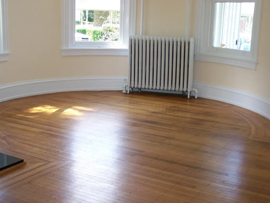 An empty room with hardwood floors, a white radiator and two sunlit windows
