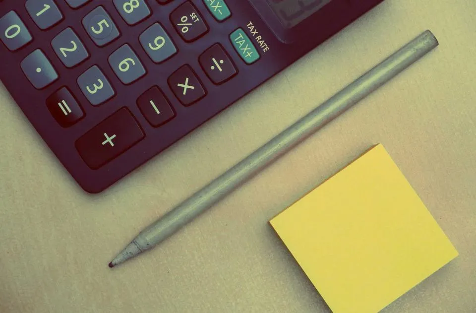 A maroon desk calculator with a tax key next to a pencil and a yellow sticky note