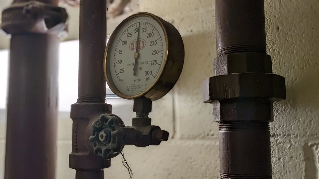 A round pressure gauge and valves on dark metal water pipes against a white wall