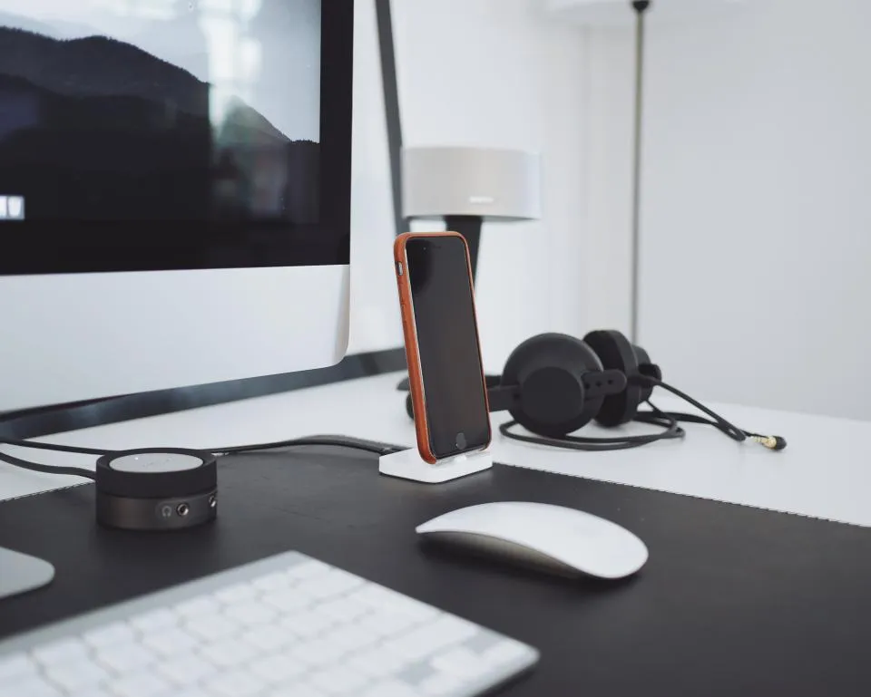 A tidy white desk with a phone on a charging stand, headphones, a mouse and a keyboard